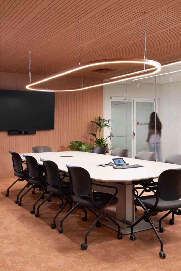 Contemporary meeting room with a white oval table, black ergonomic chairs, circular pendant lighting, wood-textured walls, and integrated screen and tech hub.