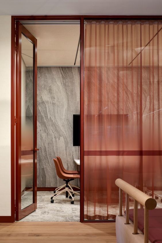 Small enclosed meeting space with a red metal-framed glass door, rust-coloured leather chair, textured grey stone wall, red sheer curtain, and a neutral carpet.