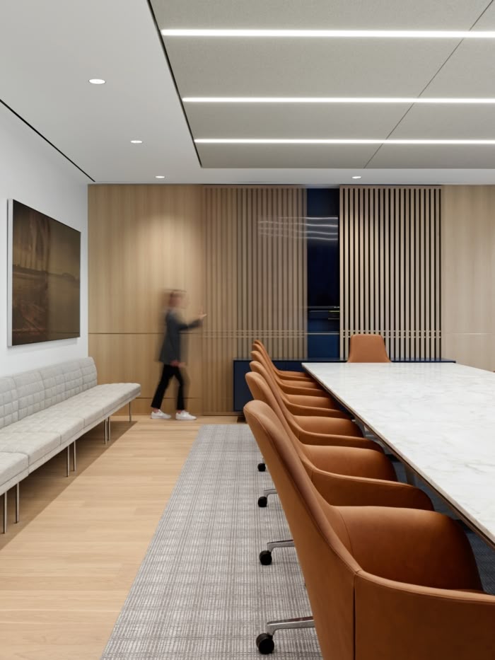 Modern boardroom with tan leather chairs around a long marble table, light wood panel walls, slatted wooden accents, and neutral tones with integrated ceiling lighting.