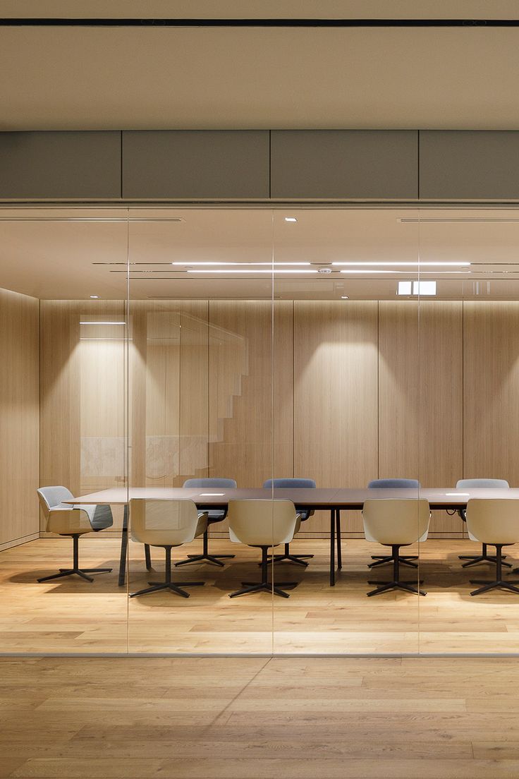 Minimalist meeting room with light wooden flooring and walls, glass enclosure, soft upholstered cream and blue chairs around a dark table, and diffused ceiling lighting.