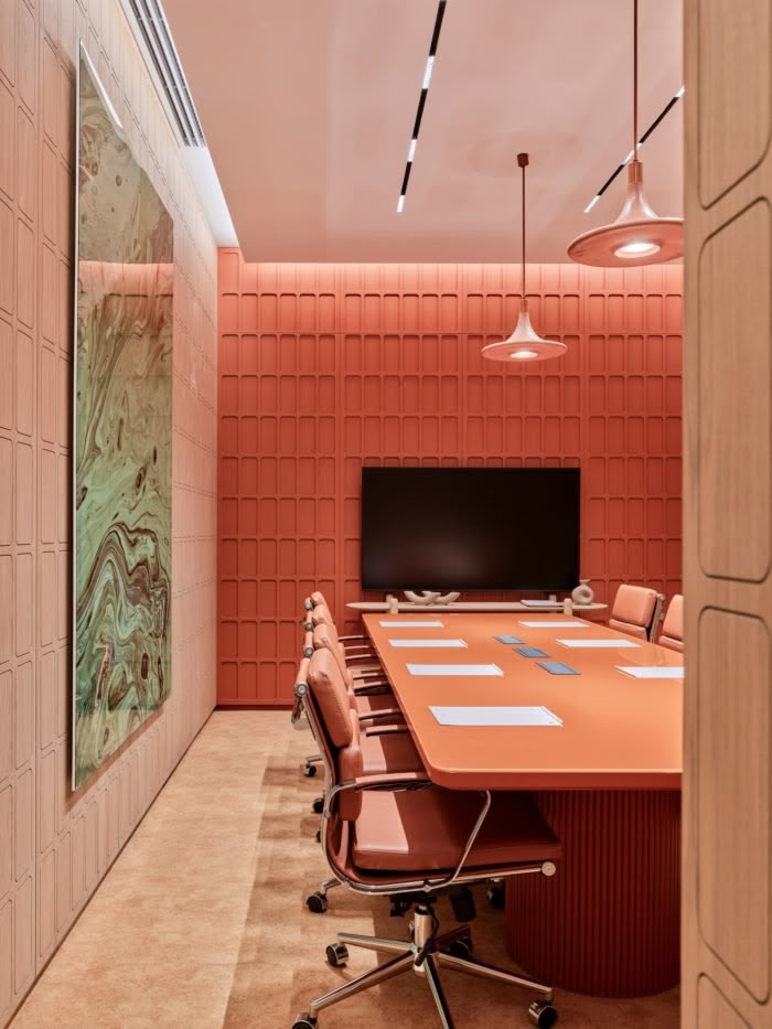 Vibrant meeting room in coral tones, featuring a matching table and chairs, textured wall panels, modern pendant lights, and a large screen mounted on the back wall.